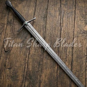 May include: A long, silver sword with a patterned blade and a black handle. The sword has a crossguard and a rounded pommel. The text "Titan Sharp Blades" is visible on the wooden surface.