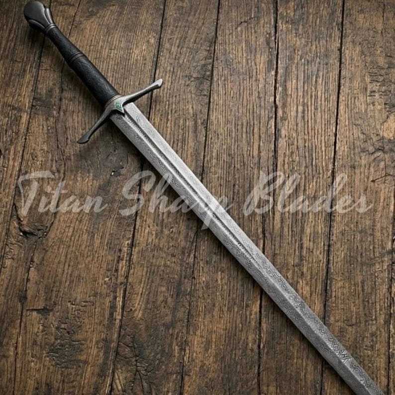 May include: A long, silver sword with a patterned blade and a black handle. The sword has a crossguard and a rounded pommel. The text "Titan Sharp Blades" is visible on the wooden surface.