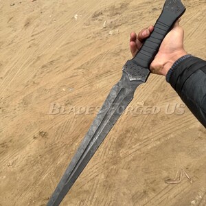 May include: A dark, long dagger with a textured, black handle and a double-edged blade. The blade has a dark, metallic finish. The text "BLADES FORGED US" is visible on the blade. The dagger is held in a hand.
