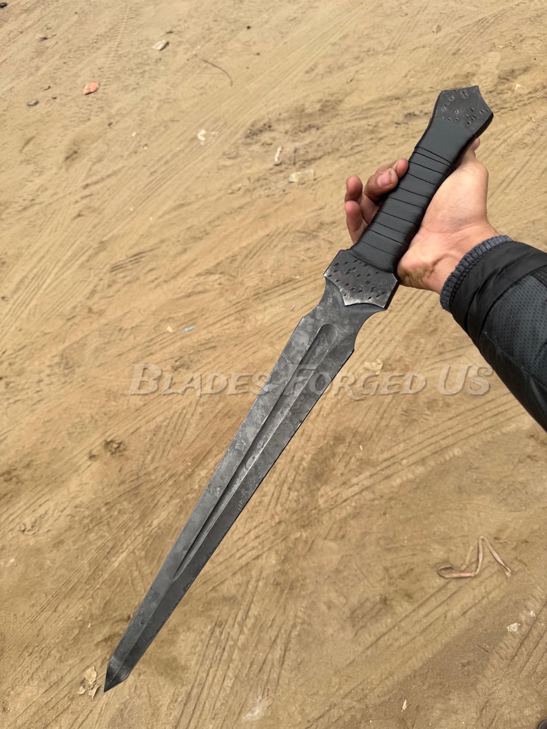 May include: A dark, long dagger with a textured, black handle and a double-edged blade. The blade has a dark, metallic finish. The text "BLADES FORGED US" is visible on the blade. The dagger is held in a hand.