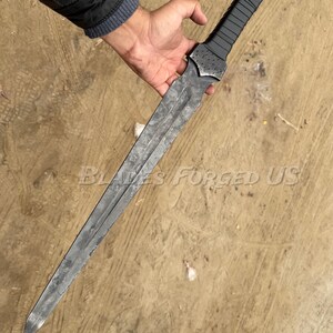 May include: A long, gray metal sword with a textured blade and a black handle. The sword is held in a person's hand. The text "Blades Forged US" is visible on the surface.