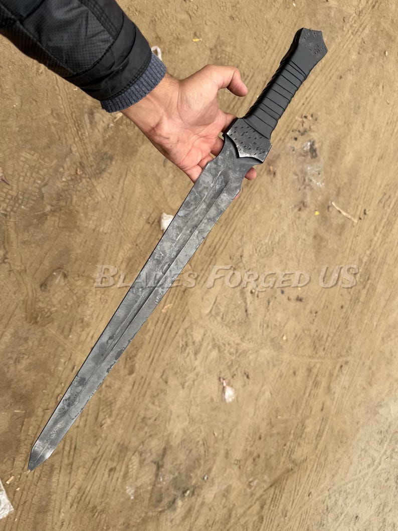 May include: A long, gray metal sword with a textured blade and a black handle. The sword is held in a person's hand. The text "Blades Forged US" is visible on the surface.