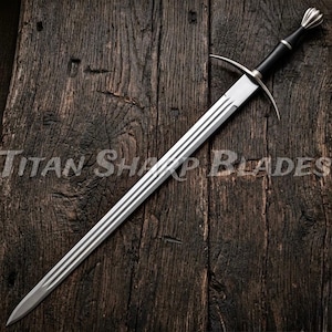 Può includere: Una lunga spada d'argento con impugnatura e guardia nere, appoggiata su una superficie di legno scuro. La lama presenta tre scanalature parallele. Il testo "Titan Sharp Blades" è visibile.