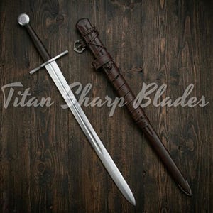 May include: A long, silver sword with a dark handle and crossguard, next to a brown leather sheath. The sword has a polished blade and a round pommel. The sheath is intricately detailed with straps and buckles. The text "Titan Sharp Blades" is visible.