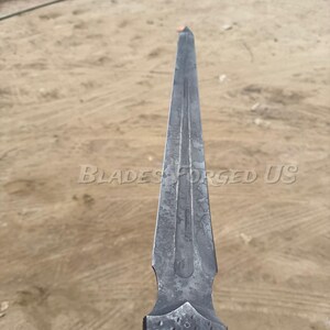 May include: A forged steel blade with a pointed tip and a textured surface. The blade has a dark gray color and features the text "Blades Forged US". The blade's design includes a central fuller and a decorative handle.