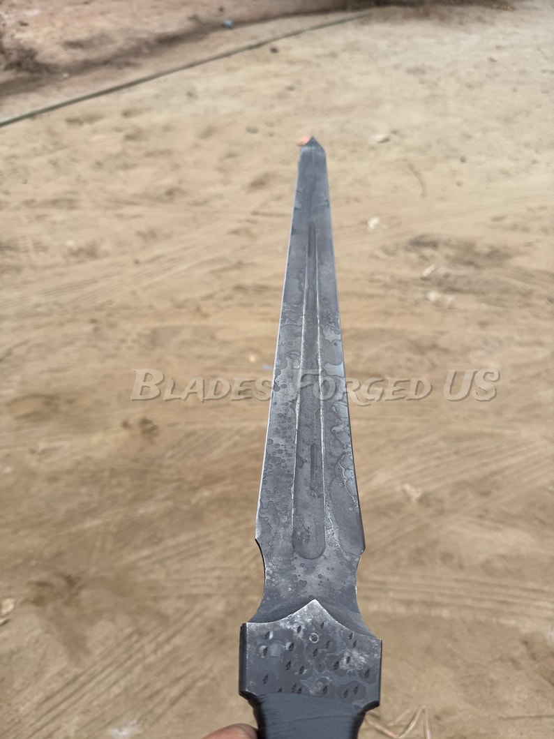 May include: A forged steel blade with a pointed tip and a textured surface. The blade has a dark gray color and features the text "Blades Forged US". The blade's design includes a central fuller and a decorative handle.
