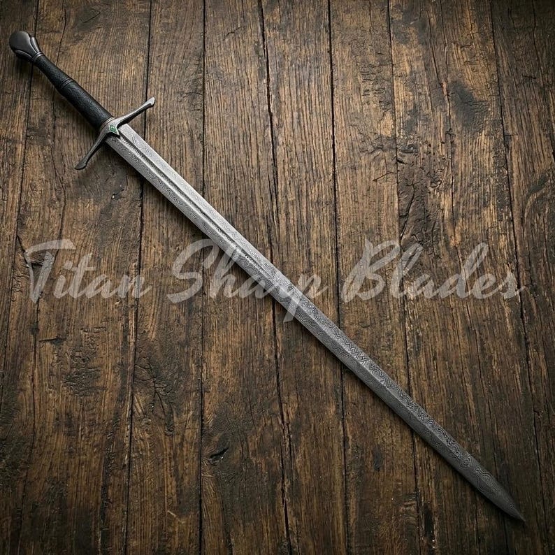 May include: A long, silver sword with a black handle and crossguard, resting on a wooden surface. The blade has a textured appearance. The text "Titan Sharp Blades" is visible on the wooden surface.