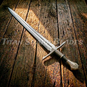 May include: A silver sword with a double-edged blade, crossguard, and ornate handle. The sword is displayed on a rustic wooden surface. The text "TITAN BLADES" is visible in the lower left corner.