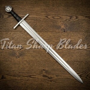 Puede incluir: Una espada de estilo medieval con una hoja plateada y un mango negro. La espada presenta una guarda cruzada y un pomo circular con un diseño. El texto "Titan Sharp Blades" es visible en la superficie de madera.