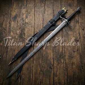 Könnte beinhalten: Ein langes, silbernes Schwert mit schwarzem Griff und Parierstange, das auf einer Holzoberfläche liegt. Das Schwert liegt neben einer schwarzen Lederscheide mit goldfarbenen Akzenten. Der Text "Titan Sharp Blades" ist sichtbar.