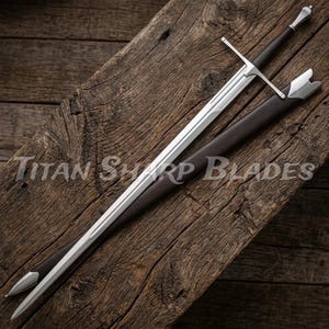 May include: A long, silver sword with a crossguard and a dark handle, resting in a brown sheath. The blade is highly polished, reflecting light. The text "Titan Sharp Blades" is visible on the wooden surface.