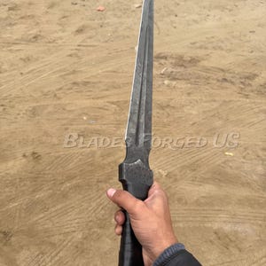 May include: A long, dark metal dagger with a pointed tip and a textured handle. The blade has a central ridge and is held in a hand. The text "BLADES FORGED US" is visible on the image.