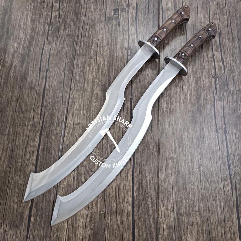 May include: Two curved swords with silver blades and brown handles are displayed on a wooden surface. The handles have a textured design with silver accents. The text "ARTISIAN SHARP CUSTOM KNIVES" is visible on the blades.