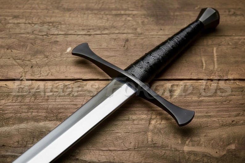 May include: A medieval-style sword with a polished steel blade and a black handle wrapped in textured material. The crossguard and pommel are also black. The sword rests on a wooden surface.