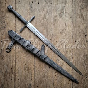 May include: A long, silver sword with a cross-shaped hilt and a rounded pommel, resting beside its gray leather scabbard. The scabbard is intricately detailed with straps and decorative elements. The sword and scabbard are set against a rustic wooden background.