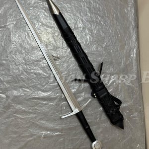 Pu&ograve; includere: Una lunga spada d'argento con un'impugnatura e una guardia nere, accanto a un fodero di cuoio nero. La spada ha una lama affilata e appuntita e un pomo d'argento. Il fodero &egrave; avvolto con cinghie di cuoio.
