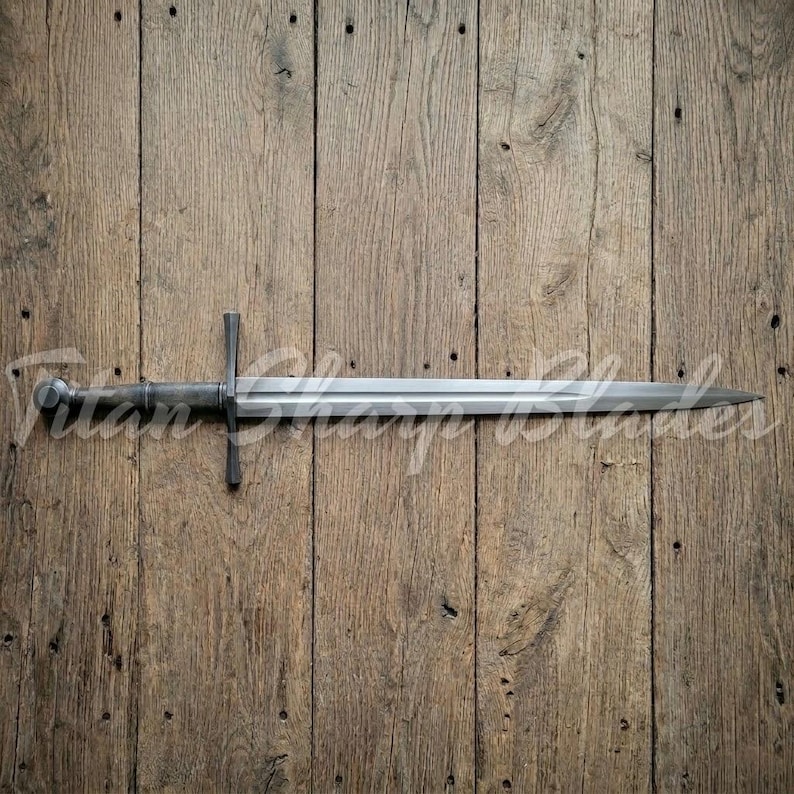Peut inclure: Une longue &eacute;p&eacute;e argent&eacute;e avec une garde en forme de croix et un pommeau arrondi. La lame est droite et se termine en pointe. L'&eacute;p&eacute;e est pr&eacute;sent&eacute;e sur un fond en bois. Le texte "Titan Sharp Blades" est visible.