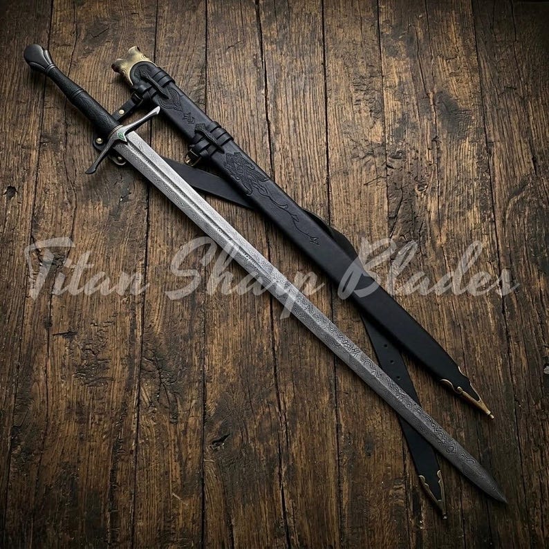 May include: A long, silver sword with a black handle and crossguard, resting on a wooden surface. The sword is accompanied by a black leather sheath with gold-colored accents. The text "Titan Sharp Blades" is visible.