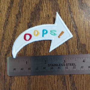 May include: White felt arrow patch with the word "OOPS!" embroidered in colorful letters. The letters are red, yellow, light blue, and green. The patch is about 3 inches long.