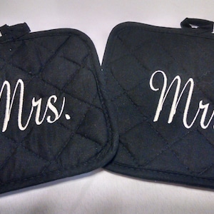 May include: Two black quilted pot holders with white embroidered text. One reads "Mrs." and the other reads "Mr.". Each pot holder has a small loop for hanging.