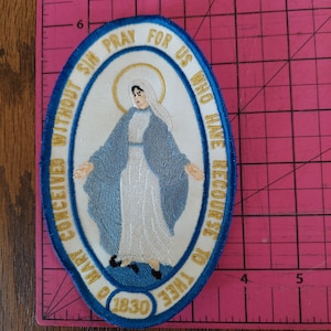 May include: Embroidered oval patch featuring a religious figure in blue and white robes, surrounded by text. The text reads "O Mary conceived without sin pray for us who have recourse to thee" and "1830". The patch has a blue border.