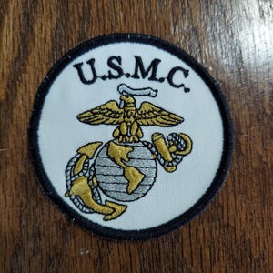 May include: A circular embroidered patch featuring a white background with a black border. The text U.S.M.C. is stitched in black at the top. Below, a gold eagle, globe, and anchor emblem is displayed with detailed gold and silver thread work.