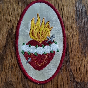 May include: A white oval embroidered patch with a red heart with a yellow flame on top. The heart is surrounded by a white wreath and has three silver swords piercing it.