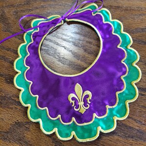 May include: A purple satin collar with a green satin trim and gold embroidery of a fleur-de-lis. The collar has a scalloped edge and a ribbon tie.