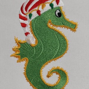 May include: Embroidered green seahorse wearing a red and white striped Santa hat with yellow and green Christmas lights. The seahorse has a yellow outline and is on a white background. This festive design is perfect for holiday decor.