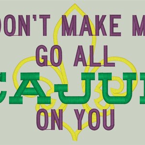 May include: Embroidered design with the text "DON'T MAKE ME GO ALL CAJUN ON YOU" in purple and green, with a yellow fleur-de-lis. The design is on a light gray background.