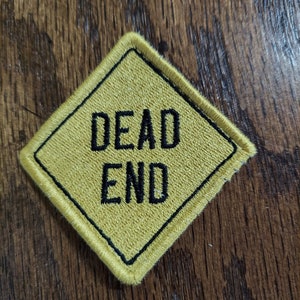 May include: A yellow diamond-shaped patch with black stitching around the edges. The patch says "DEAD END" in black embroidered letters.