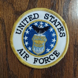 May include: A circular embroidered patch featuring the United States Air Force emblem. The design includes a central blue circle with a gold shield and black eagle, surrounded by white stars, with the text UNITED STATES AIR FORCE in black.