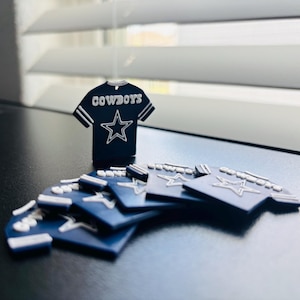 May include: A collection of small, dark blue, jersey-shaped ornaments featuring the word COWBOYS in white lettering above a white outlined star graphic. Each piece has white stripes on the sleeves and a small hole at the top for hanging purposes.