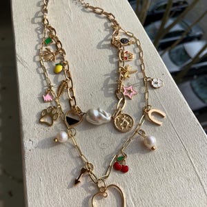 以下が含まれることがあります： ハート、馬蹄形、チェリー、足跡、星、レモン、花、鍵、トンボ、王冠、真珠、白黒の円など、複数のチャームが付いたゴールドチェーンネックレス。ネックレスには、大きな白い真珠のペンダントも付いています。