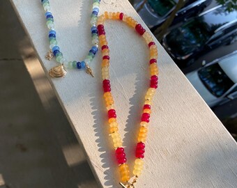 May include: Two beaded necklaces. One features blue, green, and white beads with a gold seashell charm. The other has yellow and red beads with a gold fish, starfish, and shell charm. Both necklaces are on a white surface.