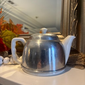 Puede incluir: Una tetera vintage con un cuerpo de metal plateado brillante y un asa, boquilla y tapa de cerámica blanca. La tetera se asienta sobre una base blanca. El asa y la boquilla son curvadas. La tapa tiene un pequeño pomo blanco.