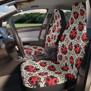 May include: A pair of car seat covers with a cream background and a repeating pattern of red ladybugs with black spots and green leaves. The seat covers are made of a soft, comfortable fabric and are easy to install.