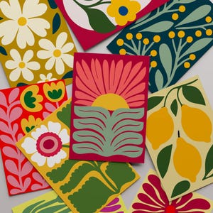 Op de afbeelding: Een verzameling kleurrijke wenskaarten met bloemen- en botanische ontwerpen. De kaarten tonen verschillende patronen, waaronder madeliefjes, citroenen en abstracte zonnestralen. Het kleurenpalet omvat tinten rood, geel, groen en blauw.