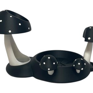 May include: Black mushroom-shaped holder with white polka dots. The holder has three sections, two of which are shaped like mushrooms. The holder is designed to hold small items, such as jewelry or other trinkets.