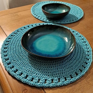 May include: Two teal crocheted placemats with a teal bowl on top of one of the placemats.