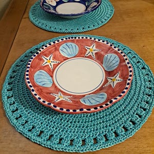 May include: Two crocheted teal placemats with white edging. One placemat has a blue and white bowl with a floral design. The other placemat has a red and white bowl with a seashell and starfish design.