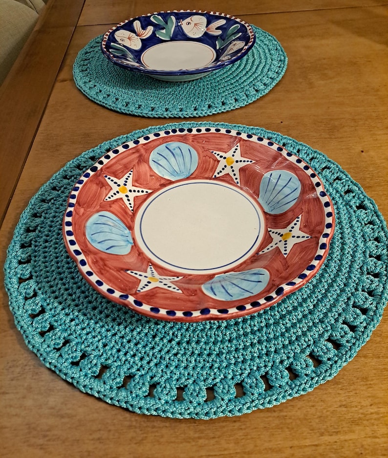 May include: Two crocheted teal placemats with white edging. One placemat has a blue and white bowl with a floral design. The other placemat has a red and white bowl with a seashell and starfish design.
