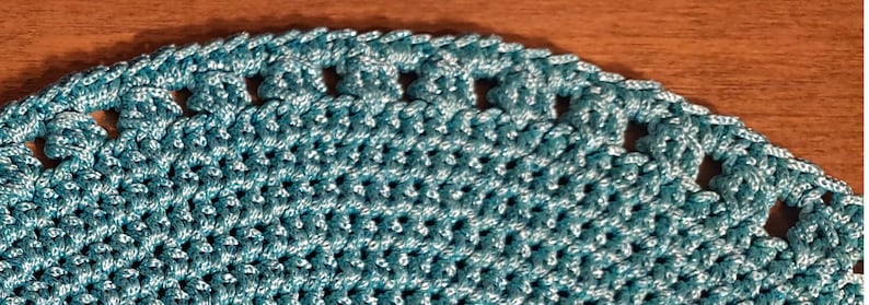 May include: A close-up of a teal crocheted doily with a scalloped edge and small holes throughout the design.