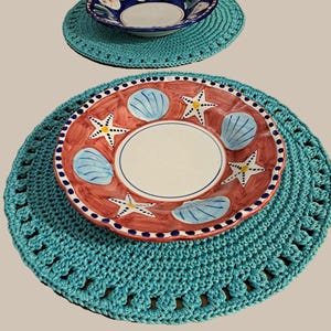 May include: Two crocheted placemats in a turquoise color with a white and blue bowl on the top placemat and a red and blue bowl with a white center on the bottom placemat.