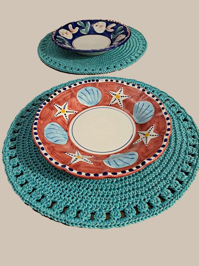 May include: Two crocheted placemats in a turquoise color with a white and blue bowl on the top placemat and a red and blue bowl with a white center on the bottom placemat.
