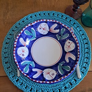 May include: A turquoise crochet placemat with a white ceramic plate featuring a blue background and a design of colorful fish.