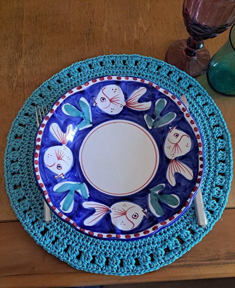 May include: A turquoise crochet placemat with a white ceramic plate featuring a blue background and a design of colorful fish.