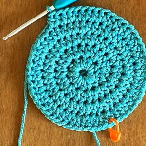 May include: A turquoise crocheted circle with a blue crochet hook and a bright orange stitch marker.