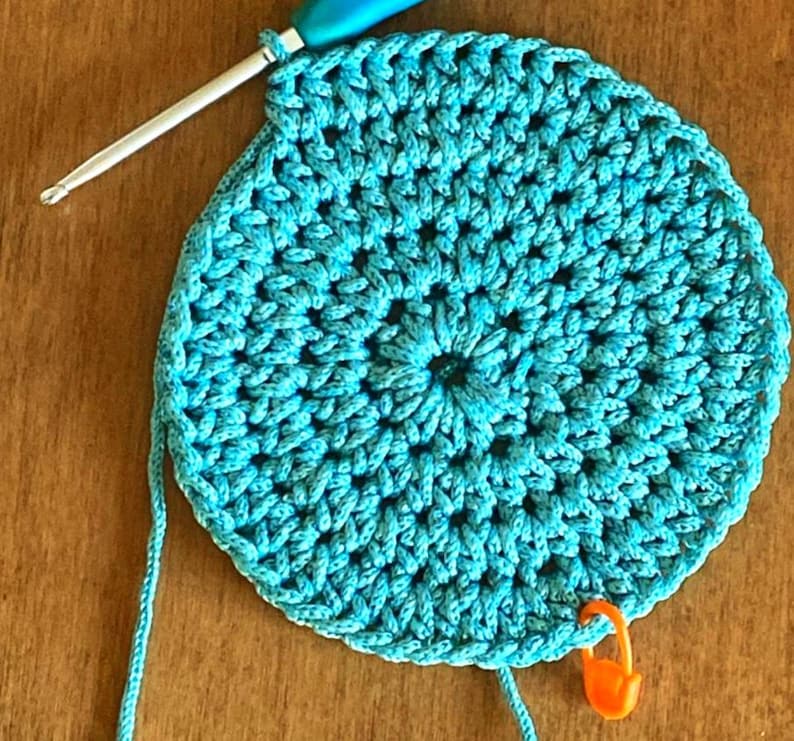 May include: A turquoise crocheted circle with a blue crochet hook and a bright orange stitch marker.