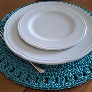 May include: A teal crocheted placemat with a white dinner plate and a smaller white plate on top. A silver fork and knife are placed on the placemat.
